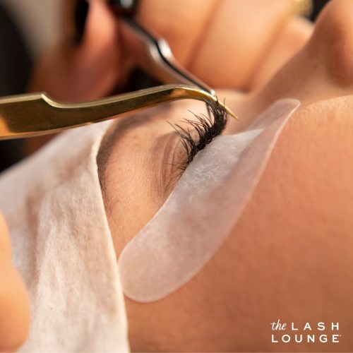 side closeup view of one closed eye of woman during a lash extension application with tweezers applying the lash extension to her natural eyelash
